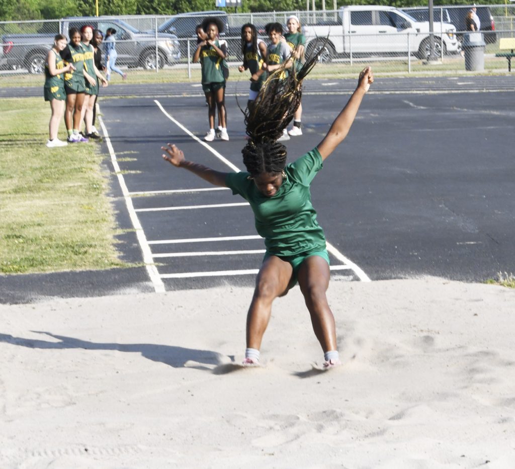 A young athlete in green lands in a sandpit during a long jump event while teammates watch in the background.