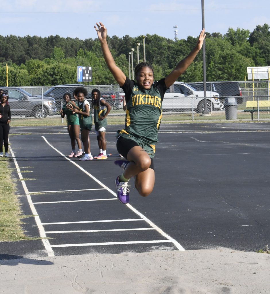 A female athlete wearing a "Vikings" jersey jumps into a sandpit during a track and field event, with teammates watching in the background.