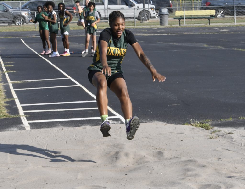 A female athlete in a "Vikings" uniform is mid-air during a long jump at a track event, with teammates watching in the background.