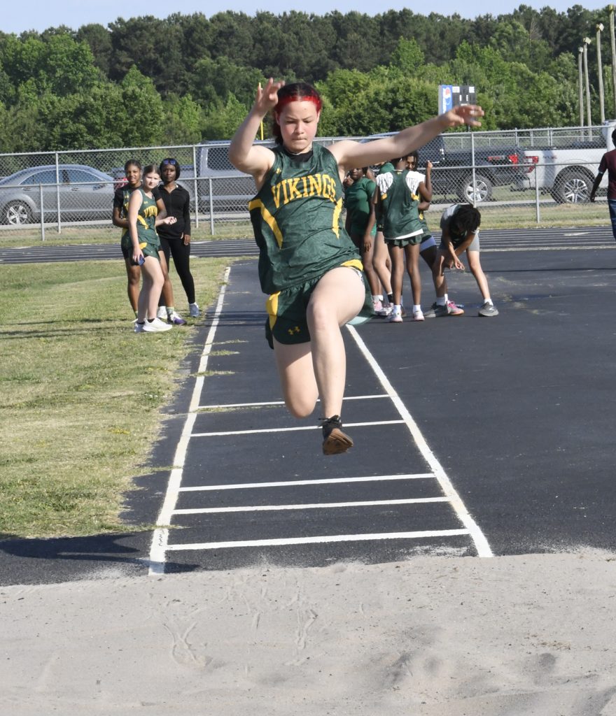A student athlete wearing a green "Vikings" uniform mid-jump during a long jump event at a school track meet.