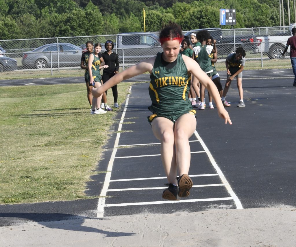 A student in a green Vikings uniform performs a long jump during a track and field event, with teammates watching in the background.