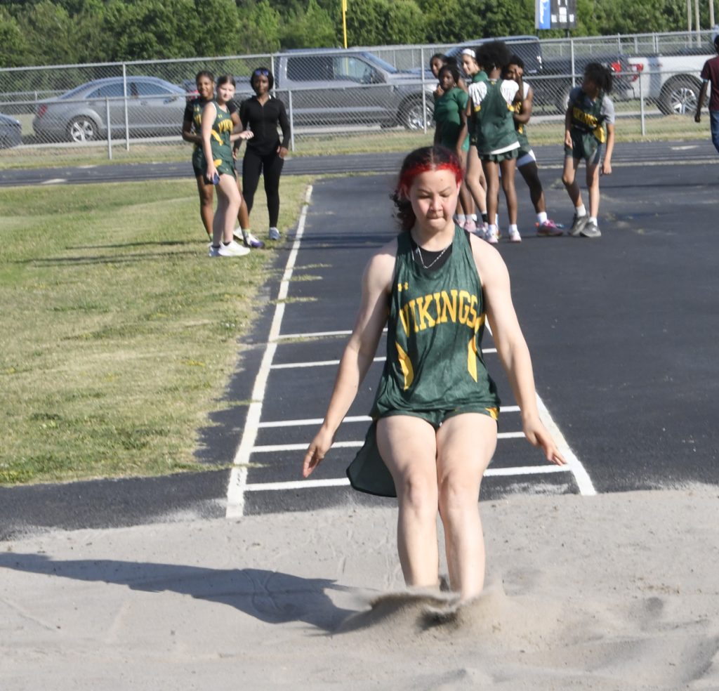 A track athlete in a green "Vikings" uniform lands in a sandpit during a long jump, with teammates and onlookers in the background.
