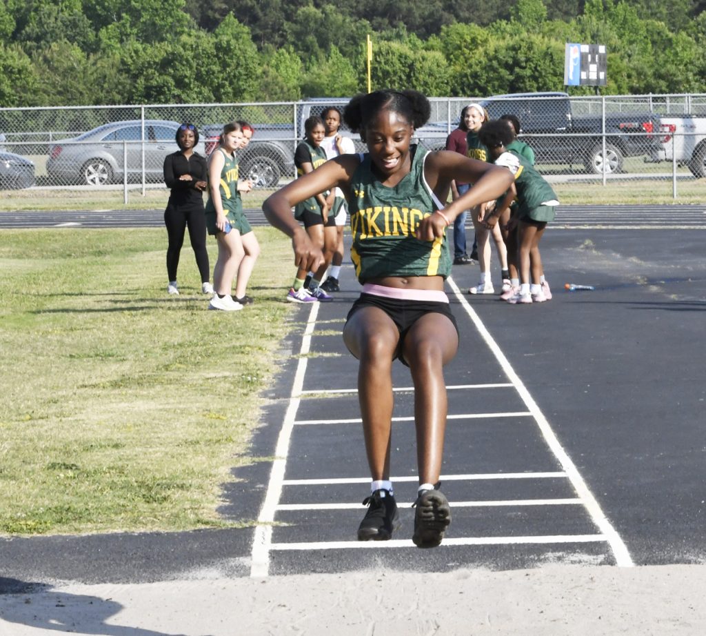 A girl in a Vikings track uniform is mid-air during a long jump, with teammates and coaches watching in the background on a sunny day.