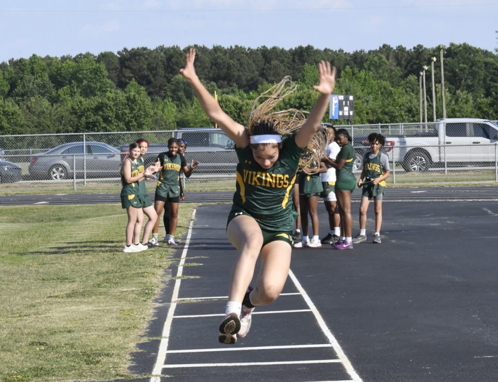 A female athlete in a green "Vikings" uniform jumps during a track and field event, with teammates cheering in the background.