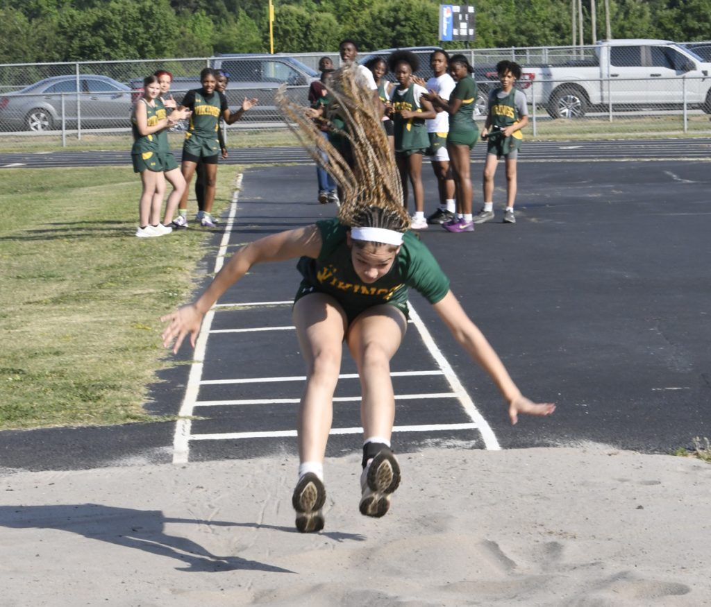 A girl in a green uniform performs a long jump at a track meet, with teammates watching and cheering in the background.