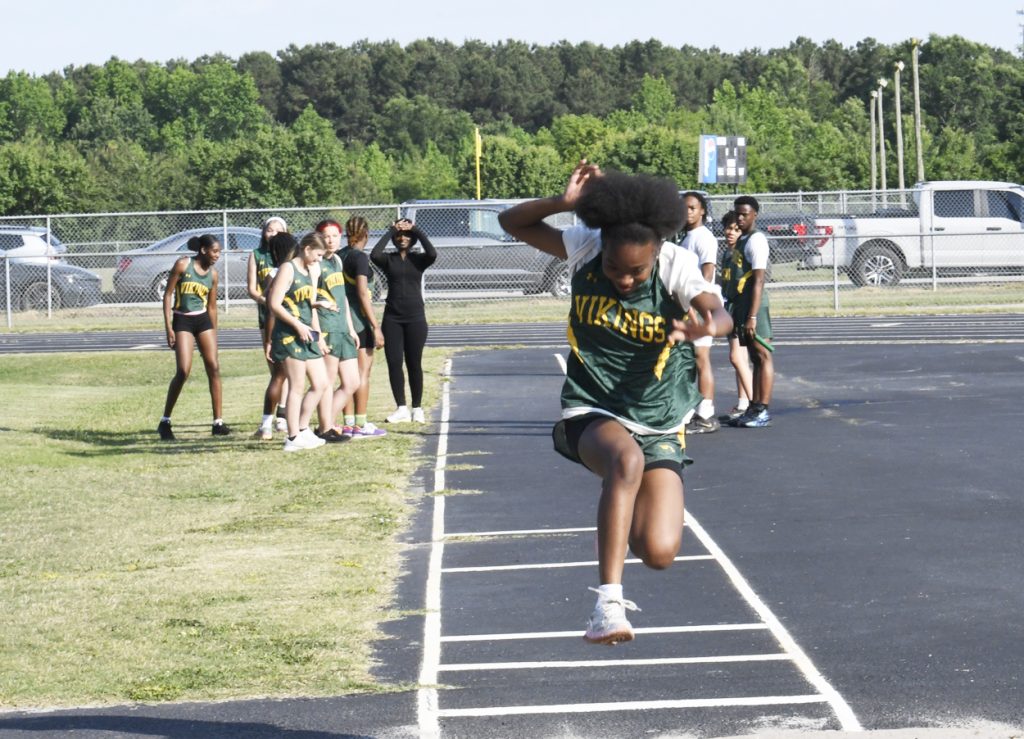 A girl in a "Vikings" uniform leaps in a track and field event, with her teammates and other students watching in the background.