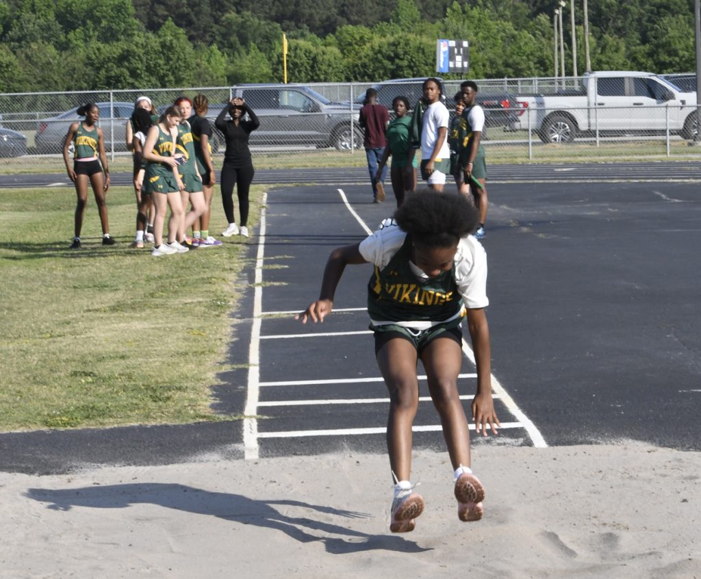 A girl in a Vikings uniform lands in a sandpit during a track and field long jump event, with teammates watching in the background.