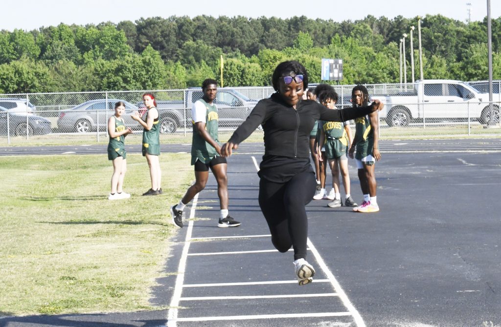 A person in black jumps on a track field during a sports event, with other athletes and students watching in the background.