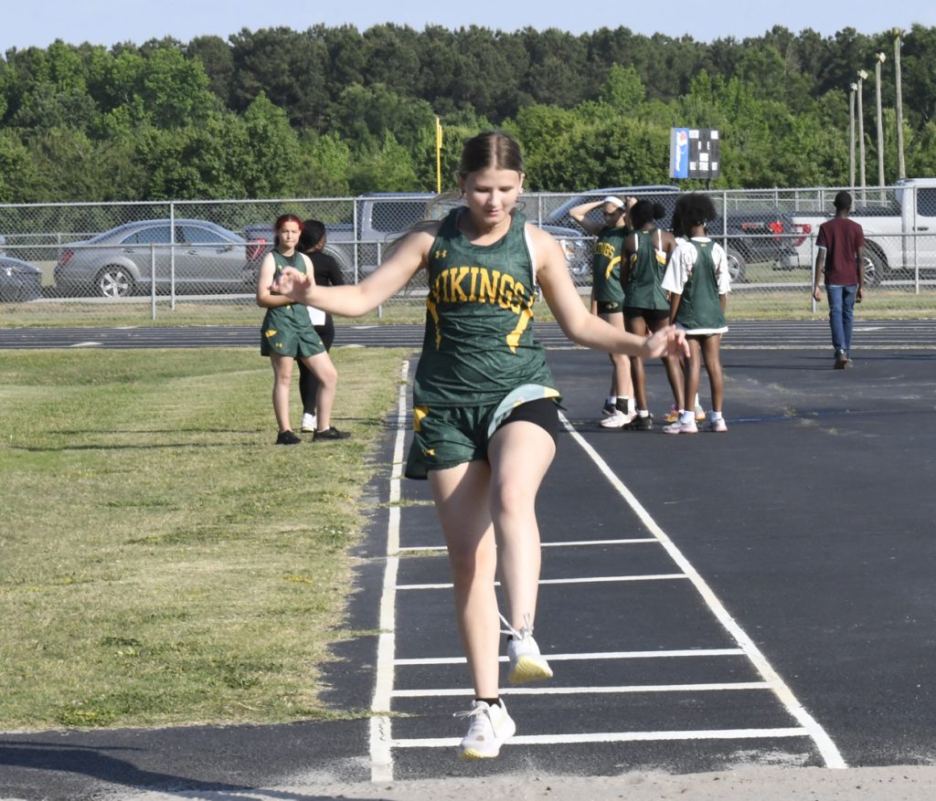 A girl in a green "Vikings" uniform jumps during a track and field event, with teammates and spectators in the background.