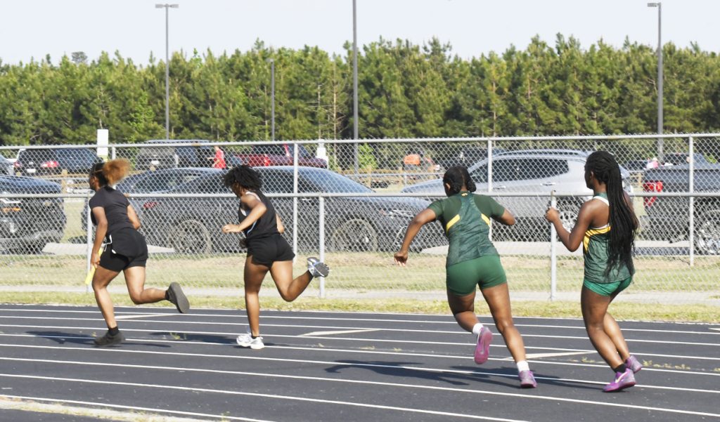 Four female athletes running a relay race on an outdoor track, preparing to pass batons, with trees and parked cars in the background.
