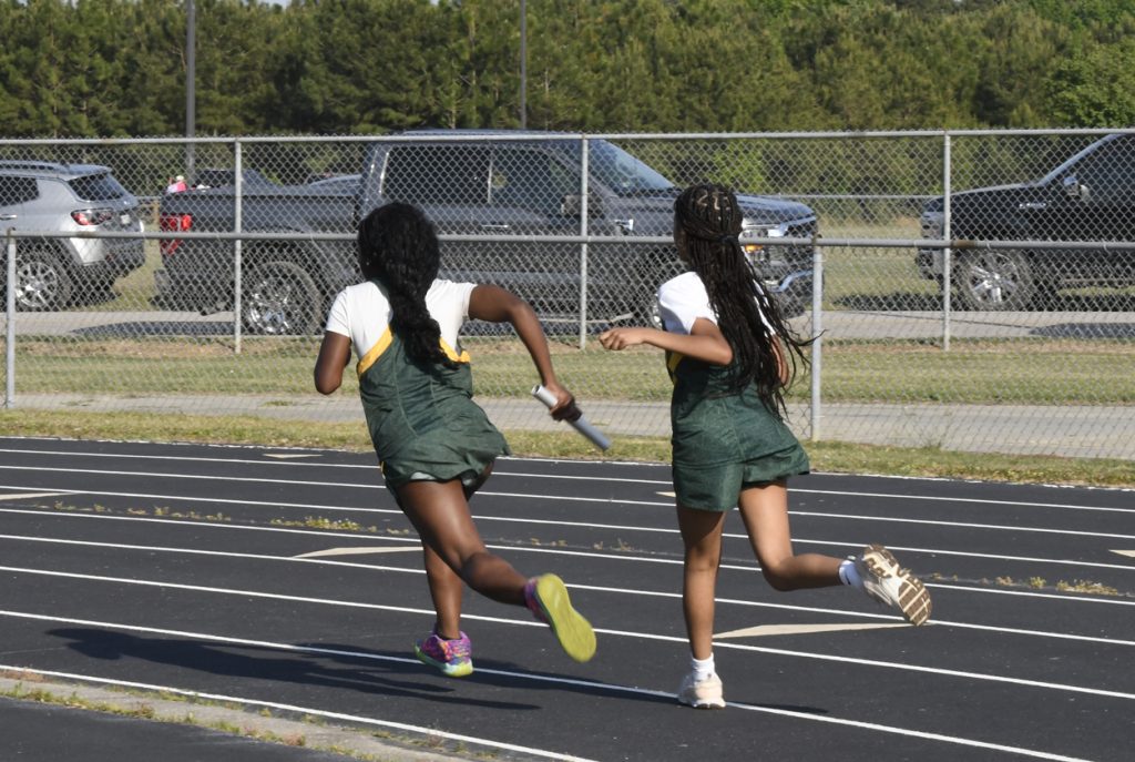 Two girls in green uniforms running a relay race on a track, one about to pass the baton to the other.
