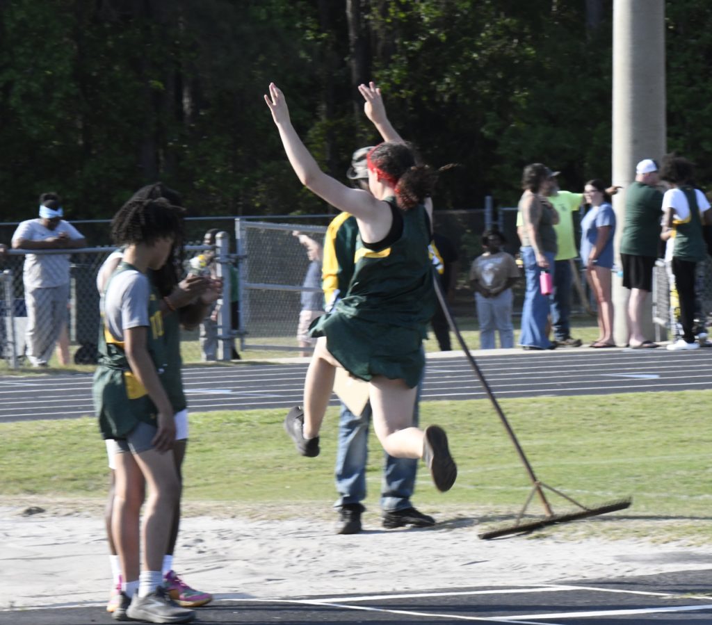 A track and field athlete jumps into a sandpit during a long jump event, with spectators and other athletes in the background.