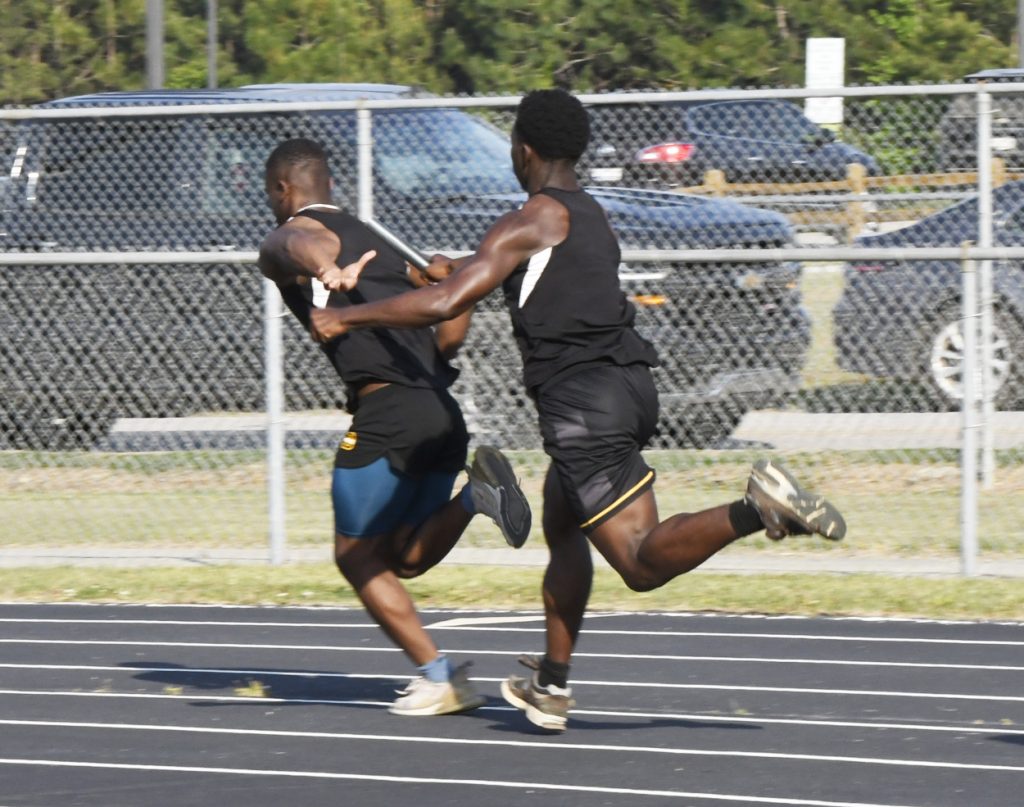 Two male athletes on a track passing a baton during a relay race, both wearing black athletic outfits, with parked cars and fence in the background.