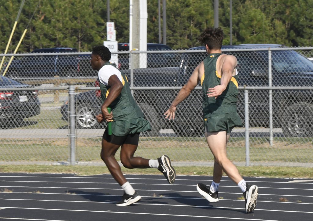 Two male athletes in green uniforms running on a track during a relay race, one passing the baton to the other.
