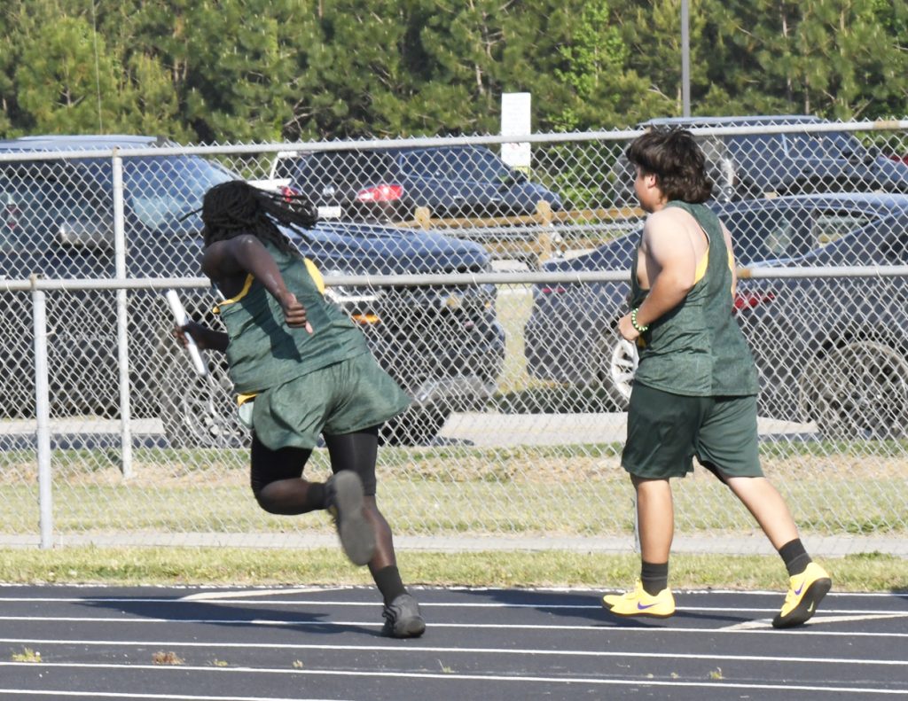 Two athletes wearing green uniforms participate in a track relay race, one running with a baton while the other walks nearby.