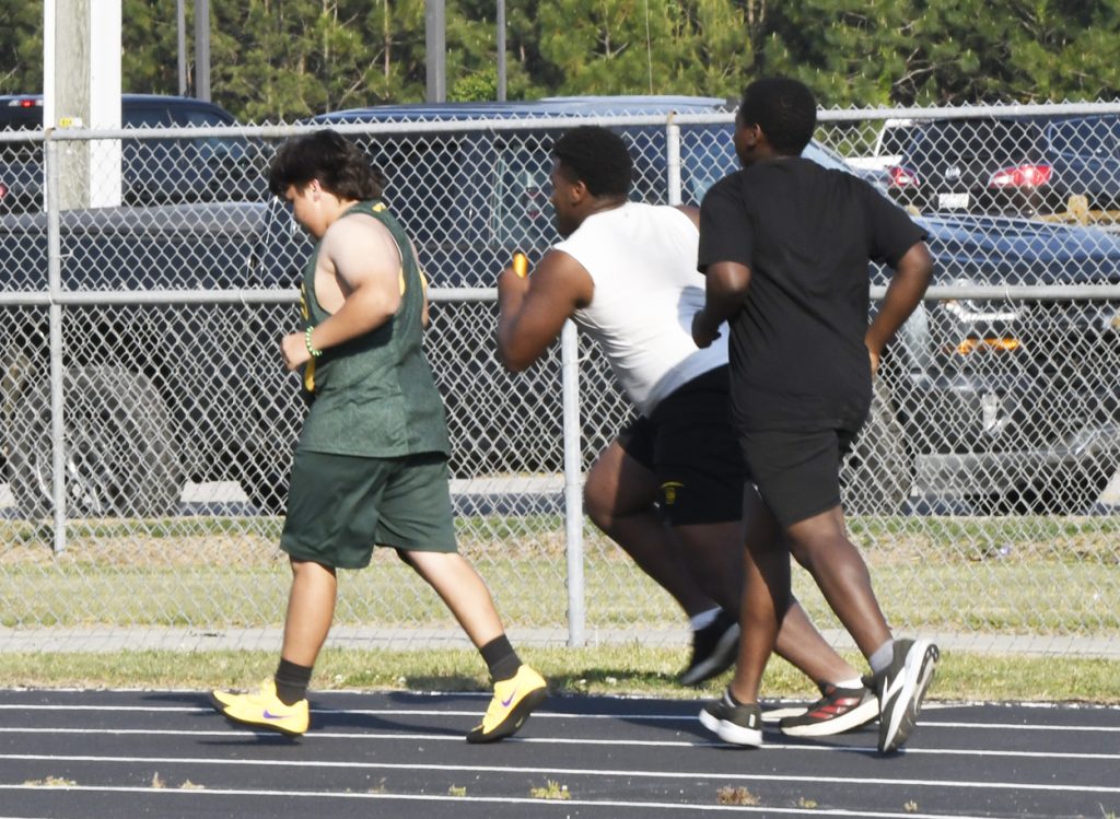 Three athletes running a relay race on a track, passing batons, with a chain-link fence and parked vehicles in the background.