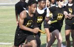 High school track team members in black uniforms pass batons during a relay race on an outdoor track, with teammates cheering in the background.