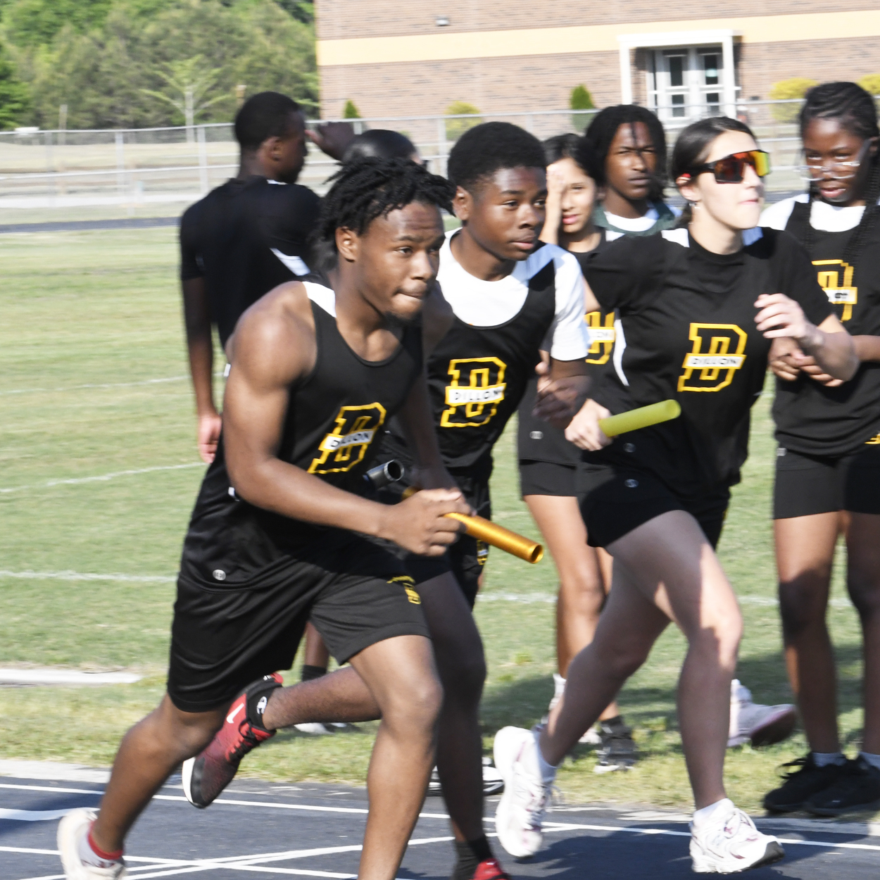High school track team members in black uniforms pass batons during a relay race on an outdoor track, with teammates cheering in the background.