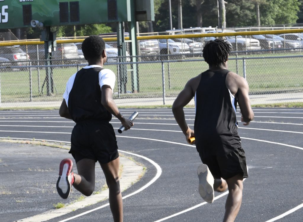Two male athletes running on a track, each holding a relay baton, during a relay race on a sunny day.
