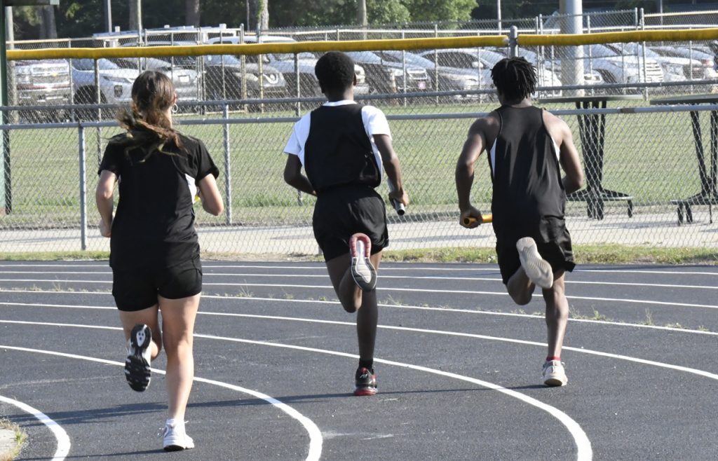 Three athletes running on a track during a relay race, holding batons, viewed from behind.