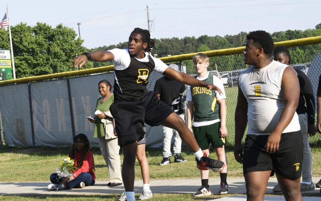 A track and field athlete in mid-action, preparing to throw a discus, with teammates and spectators watching from the sidelines near a fence.