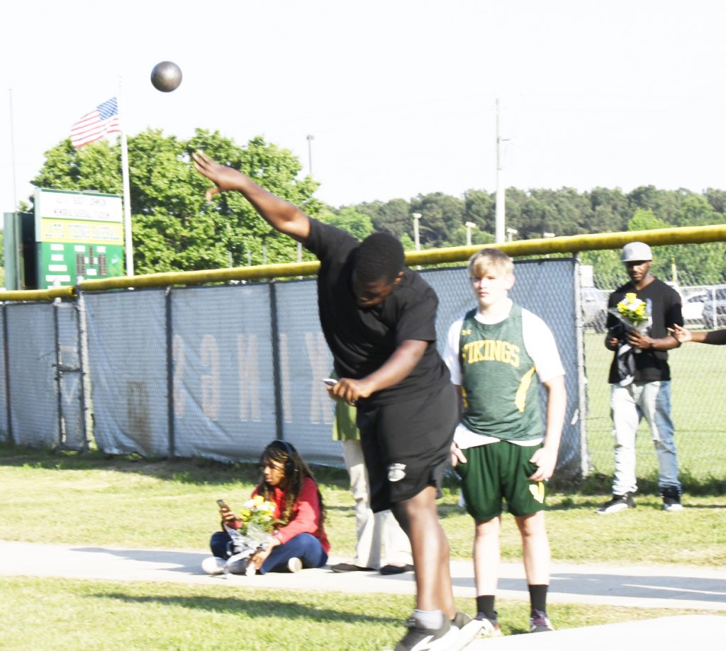 A student athlete throws a shot put during a track and field event, with spectators and other participants watching nearby on a sunny day.