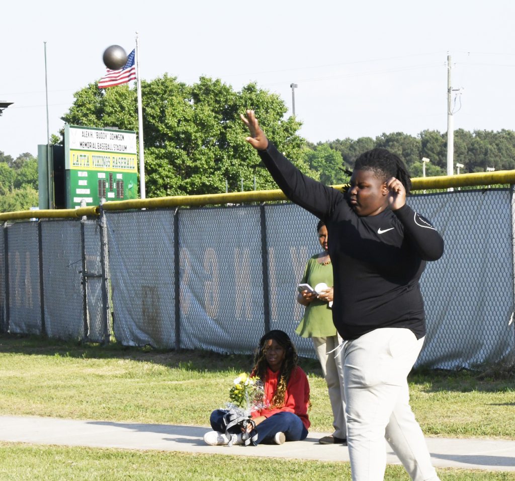 A person in athletic wear throws a shot put outdoors near a baseball field, with an American flag and scoreboard in the background, and two people nearby.