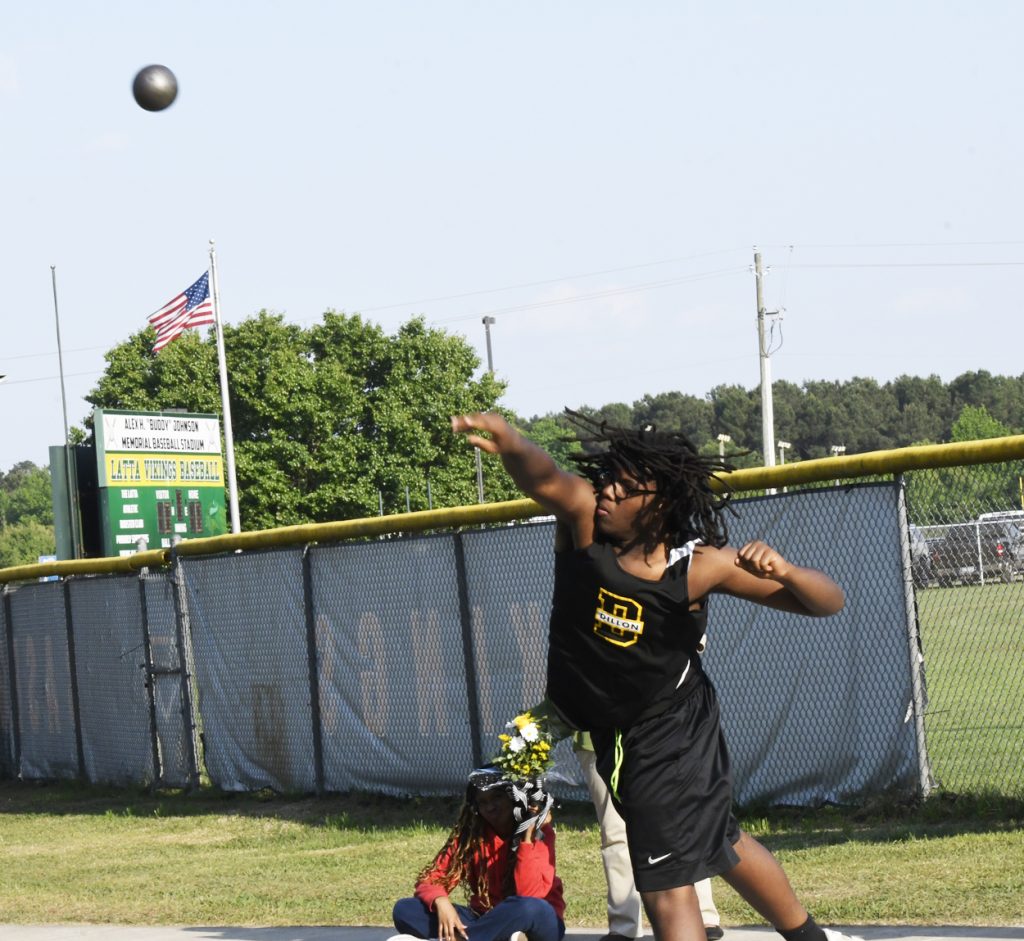 A young athlete in black sportswear throws a shot put outdoors near a baseball field, with an American flag and scoreboard visible in the background.