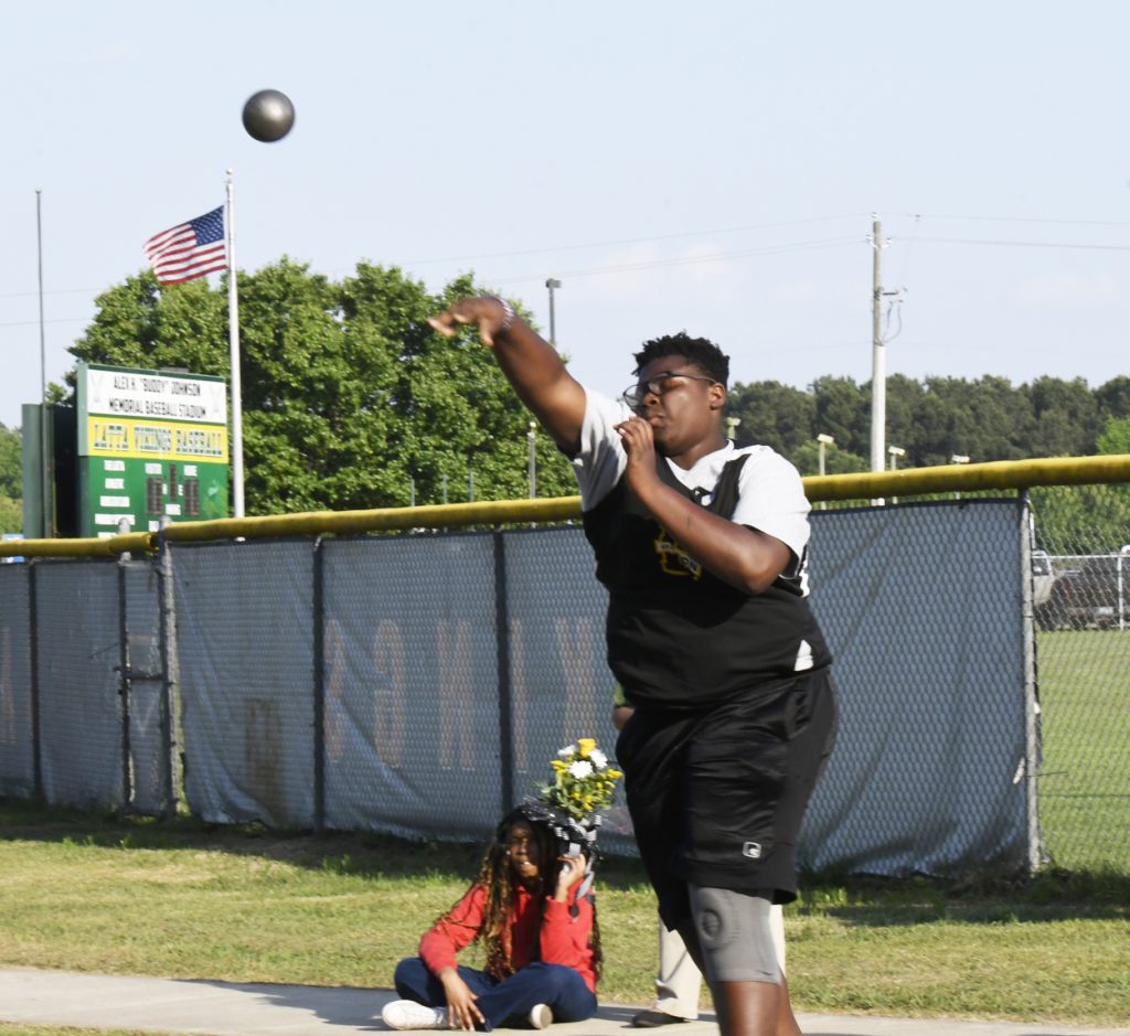 Athlete in a black uniform participates in shot put, throwing a heavy ball outdoors near a fence, with a scoreboard and American flag in the background.