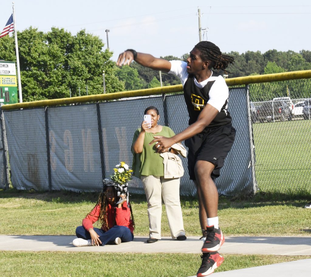 A young male athlete throws a shot put while two women watch; one films with her phone, and the other sits holding flowers.
