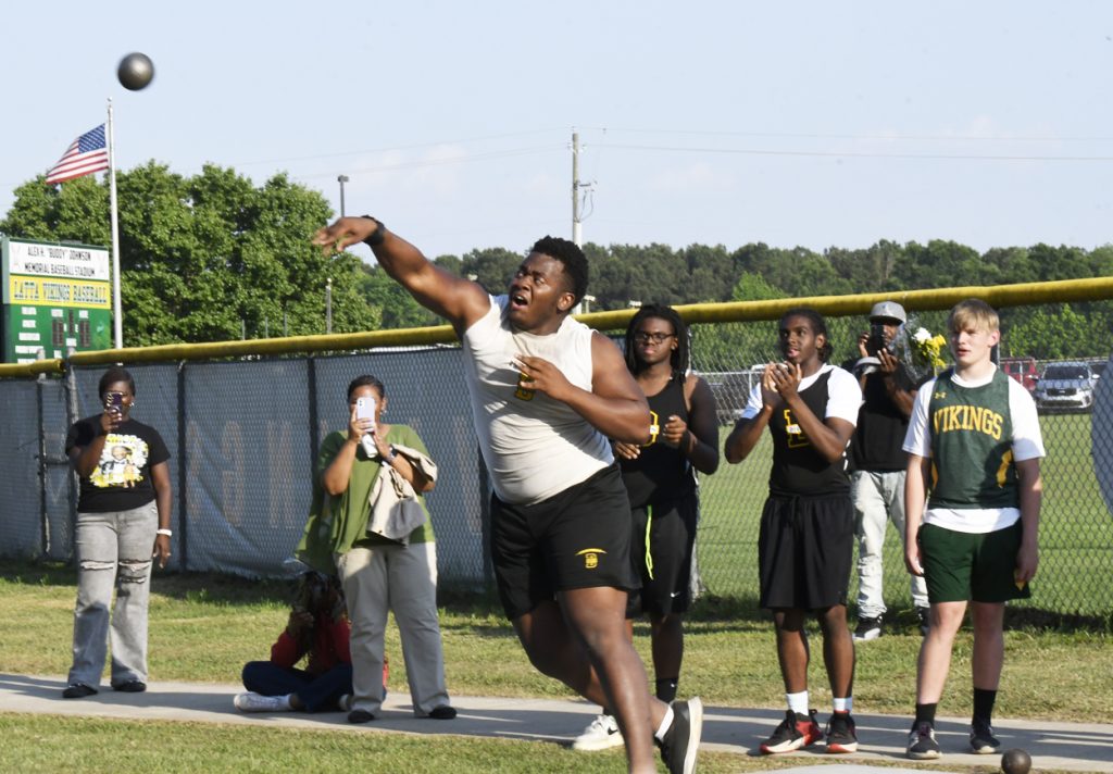 A student athlete throws a shot put during a track and field event, with spectators and other competitors watching and capturing photos in the background.