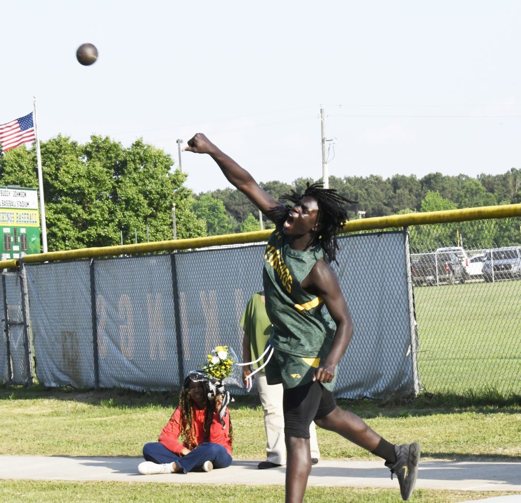 A male athlete in a green uniform throws a shot put outdoors, with a fence, scoreboard, and American flag in the background.