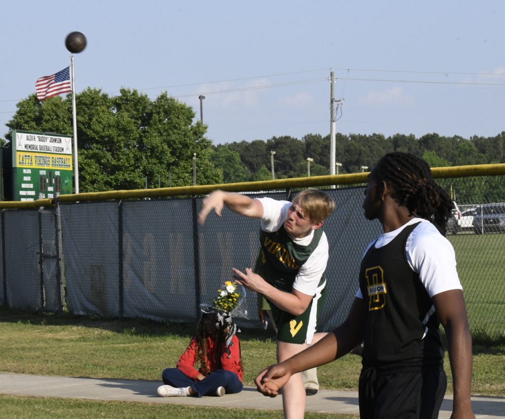 A high school athlete throws a shot put while another competitor watches during a track and field event. An American flag and scoreboard are visible in the background.