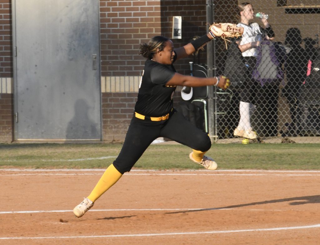 A softball pitcher in black and yellow uniform leaps forward while pitching, with players and equipment visible in the background behind a chain-link fence.