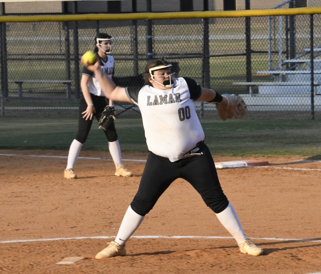 A softball pitcher in a "LAMAR" jersey prepares to throw the ball, with another player ready in the background on a dirt field.