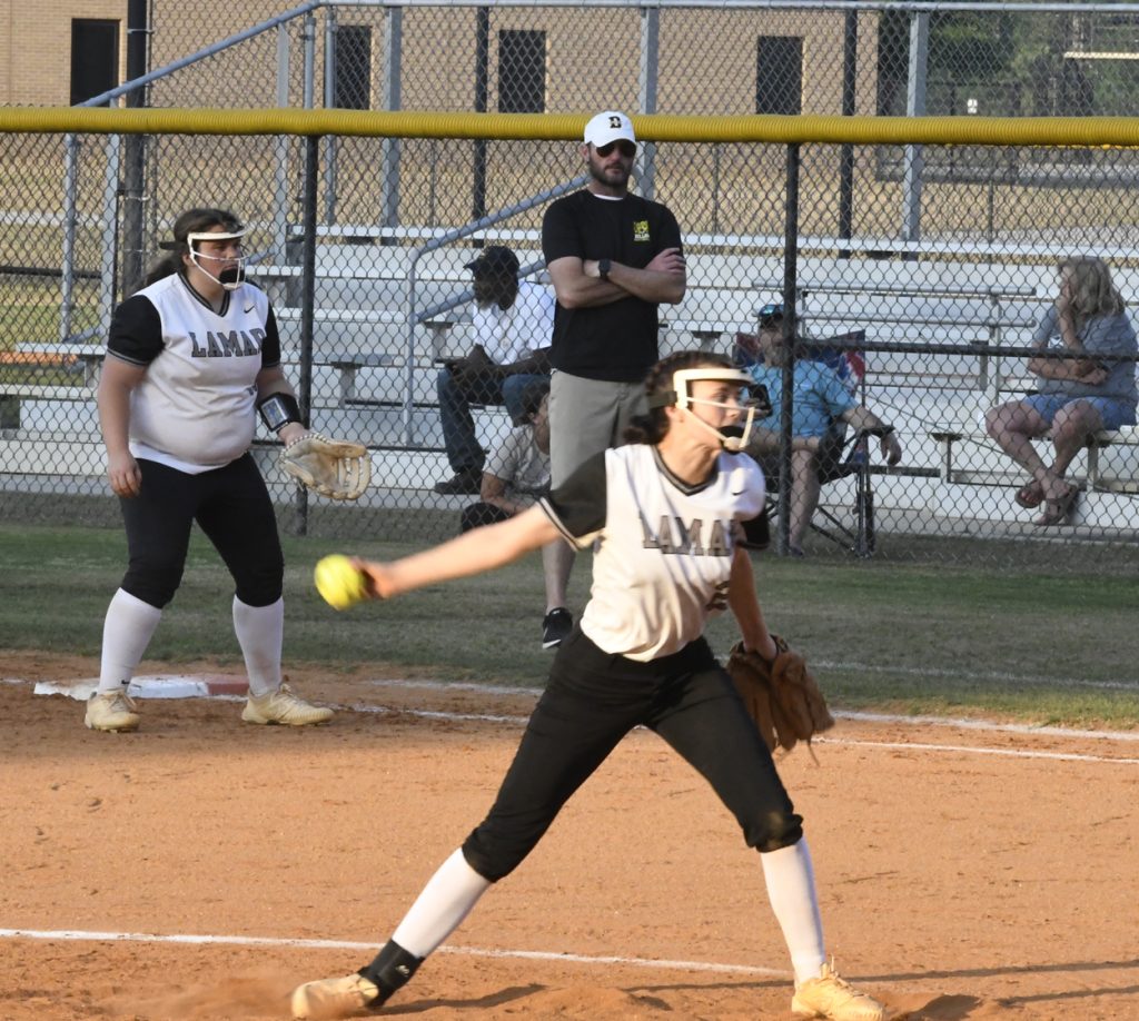 A softball pitcher in mid-throw with a teammate and coach watching, both wearing "LAMAR" uniforms, on a dirt field with a chain-link fence and spectators in the background.