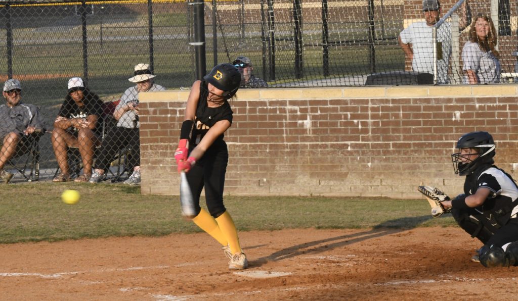 A softball player in black uniform hits a yellow ball during a game, while a catcher and spectators watch in the background.