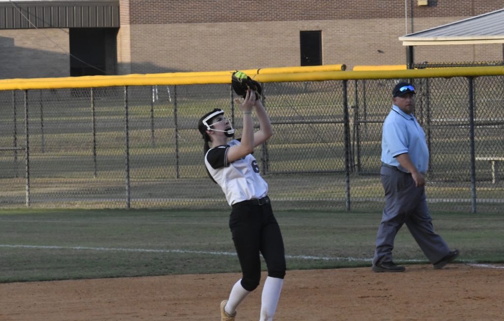 A softball player catches a ball above her head on the field as an umpire walks nearby.