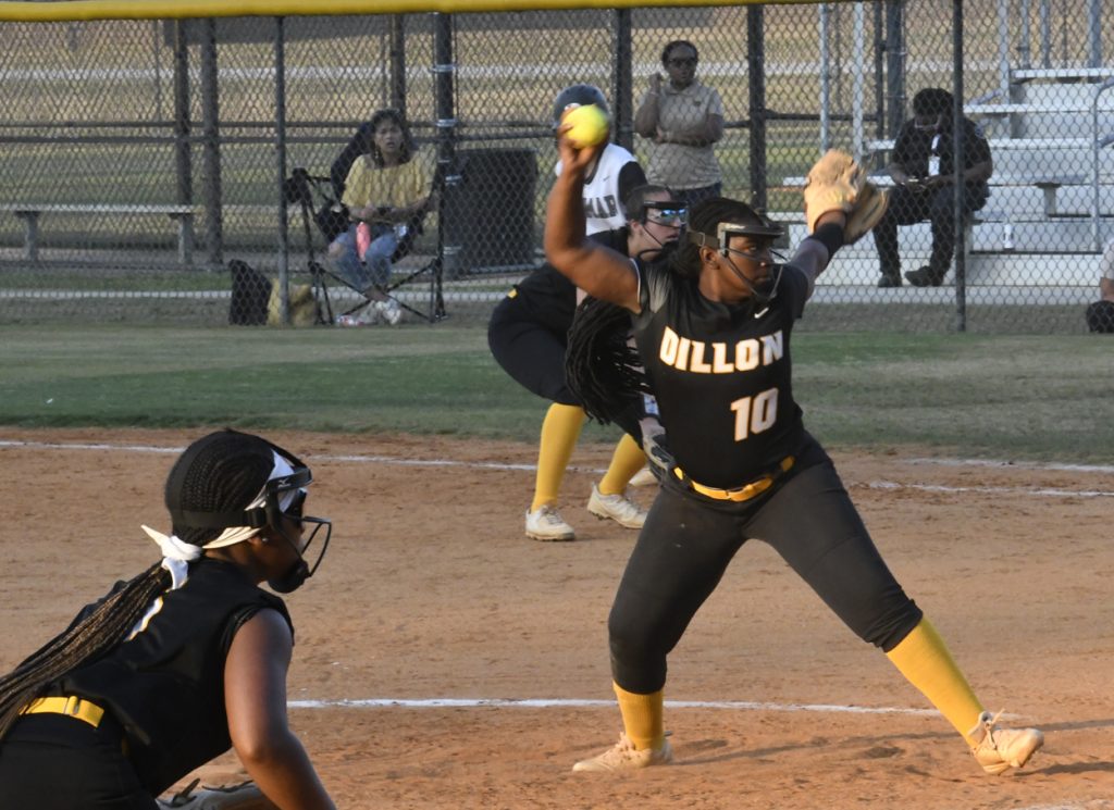 A softball pitcher in a black “Dillon” jersey throws the ball mid-game, while a catcher and teammates are ready on the field, with spectators in the background.