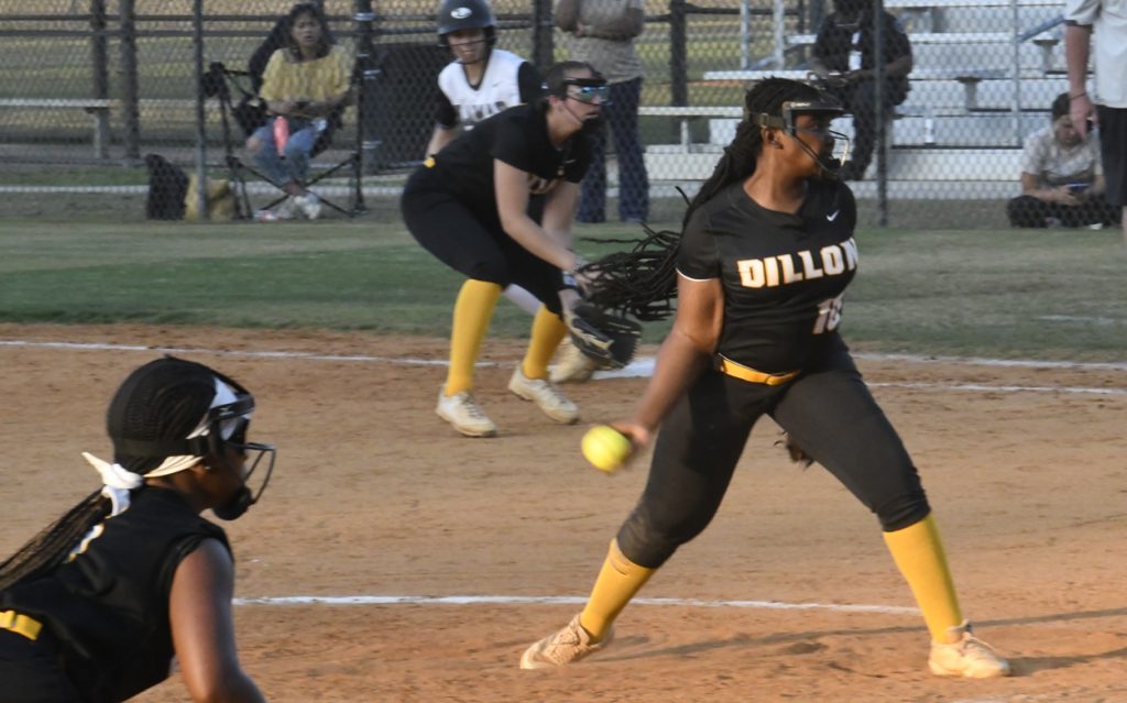 A softball pitcher in black uniform mid-pitch, with teammates ready in the field, playing on a dirt and grass field, spectators visible behind fence.