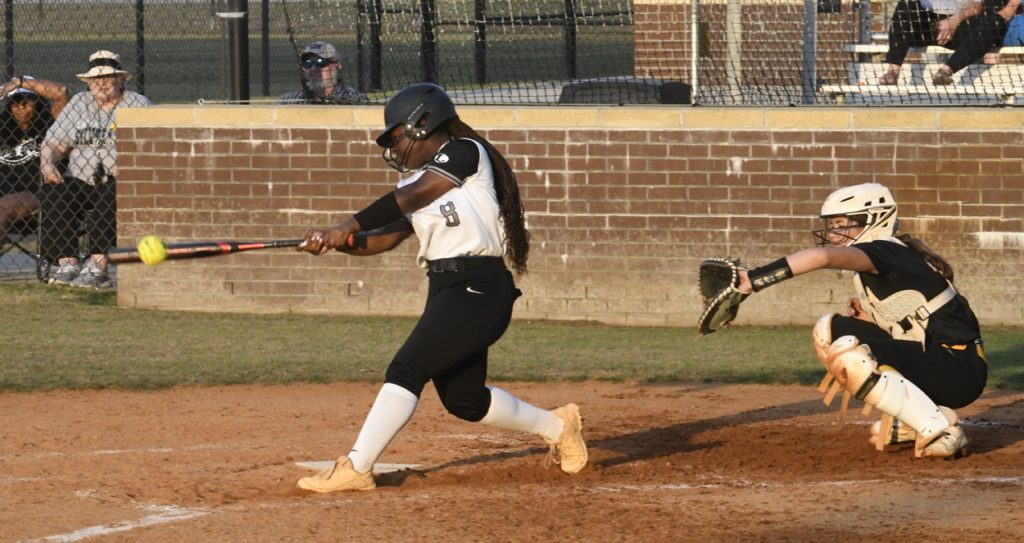 A softball player in a white and black uniform swings at a pitch while the catcher is ready behind home plate. Spectators watch from the stands.