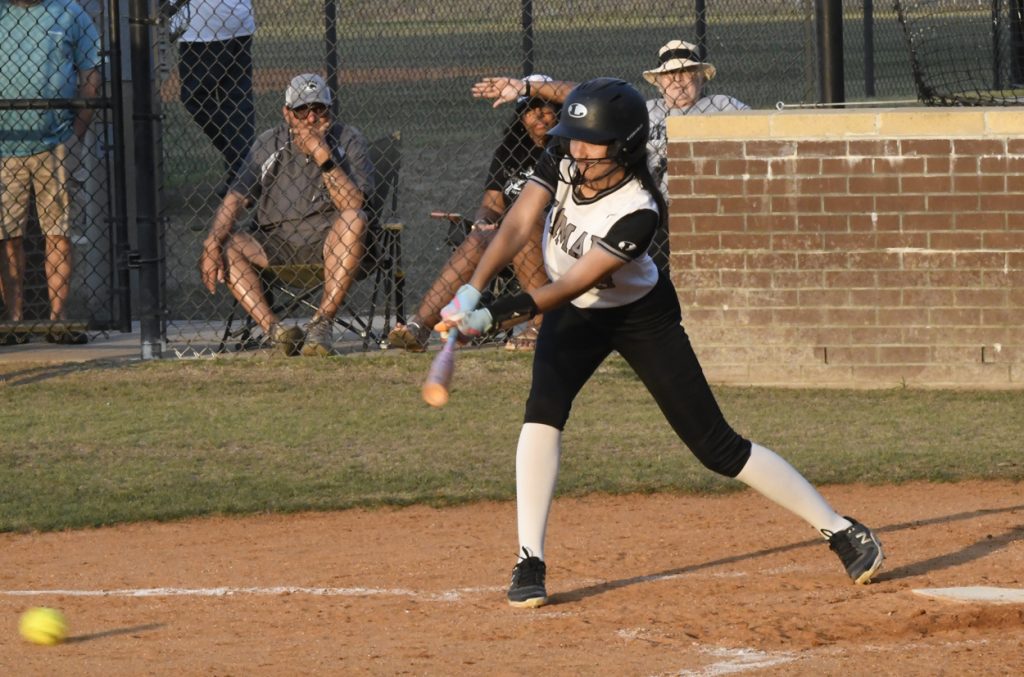 A young softball player in uniform swings at a pitched ball during a game, with spectators watching from behind a chain-link fence.
