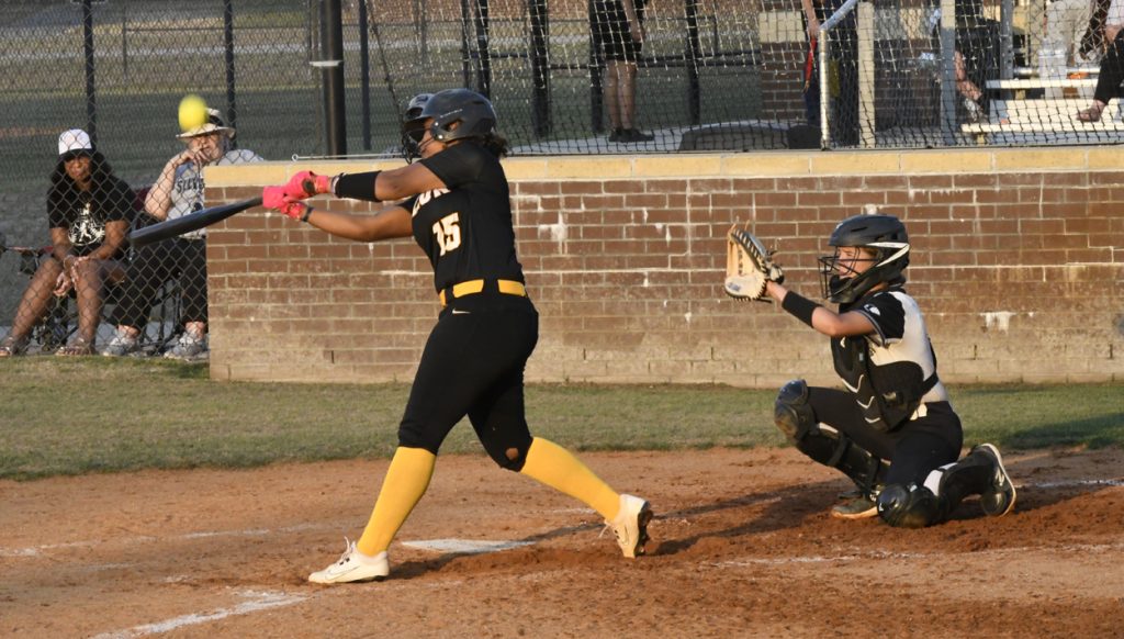 A softball player in black swings at a pitch while the catcher crouches behind home plate, ready to catch the ball. Spectators watch from behind a fence.