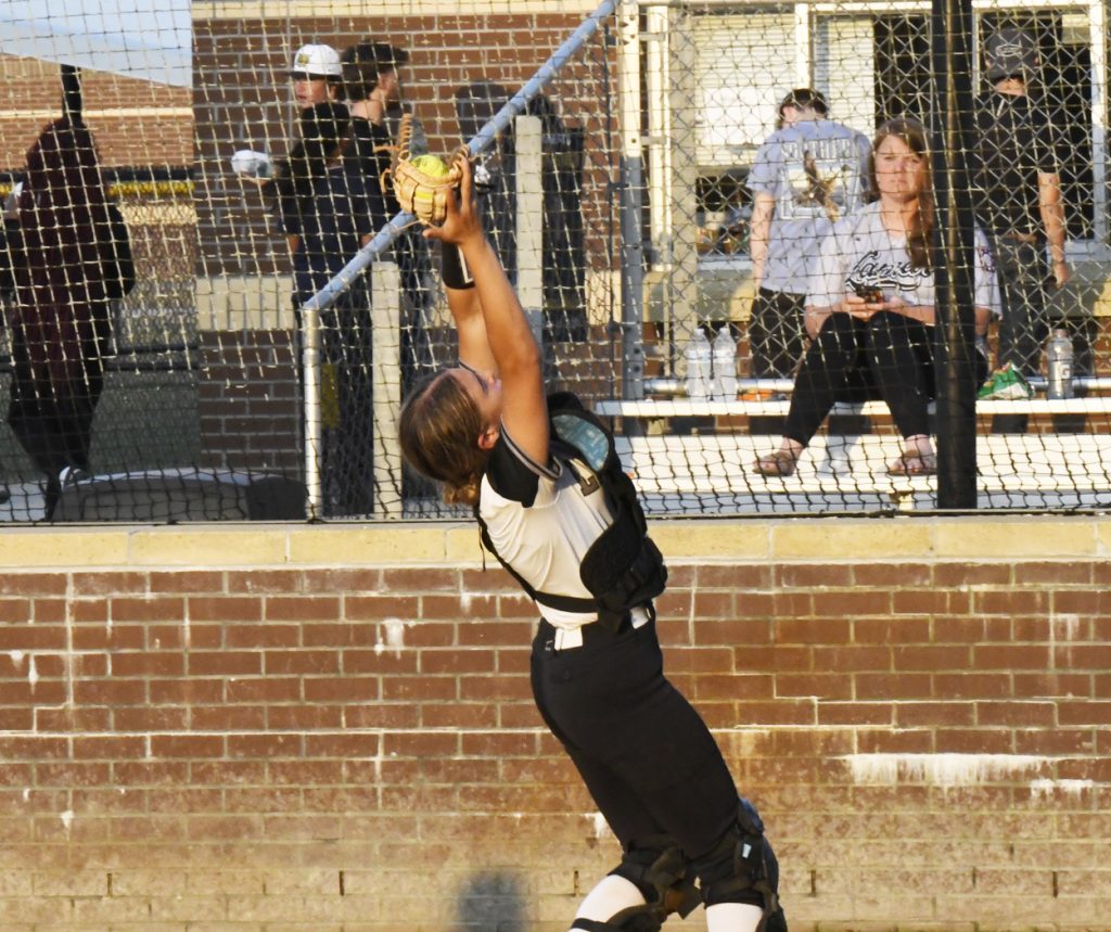 Softball catcher reaches high in full gear to catch a ball near a brick wall, with spectators and other players in the background behind a net.