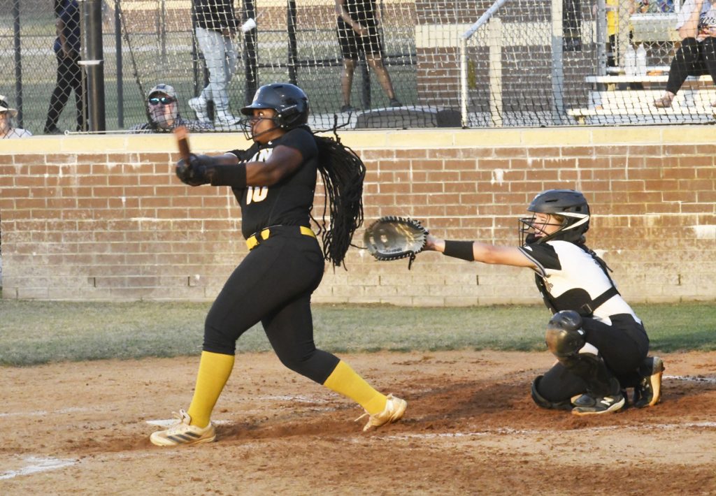 A softball player swings at a pitch while the catcher reaches out to catch the ball, during a game on a dirt field with spectators in the background.
