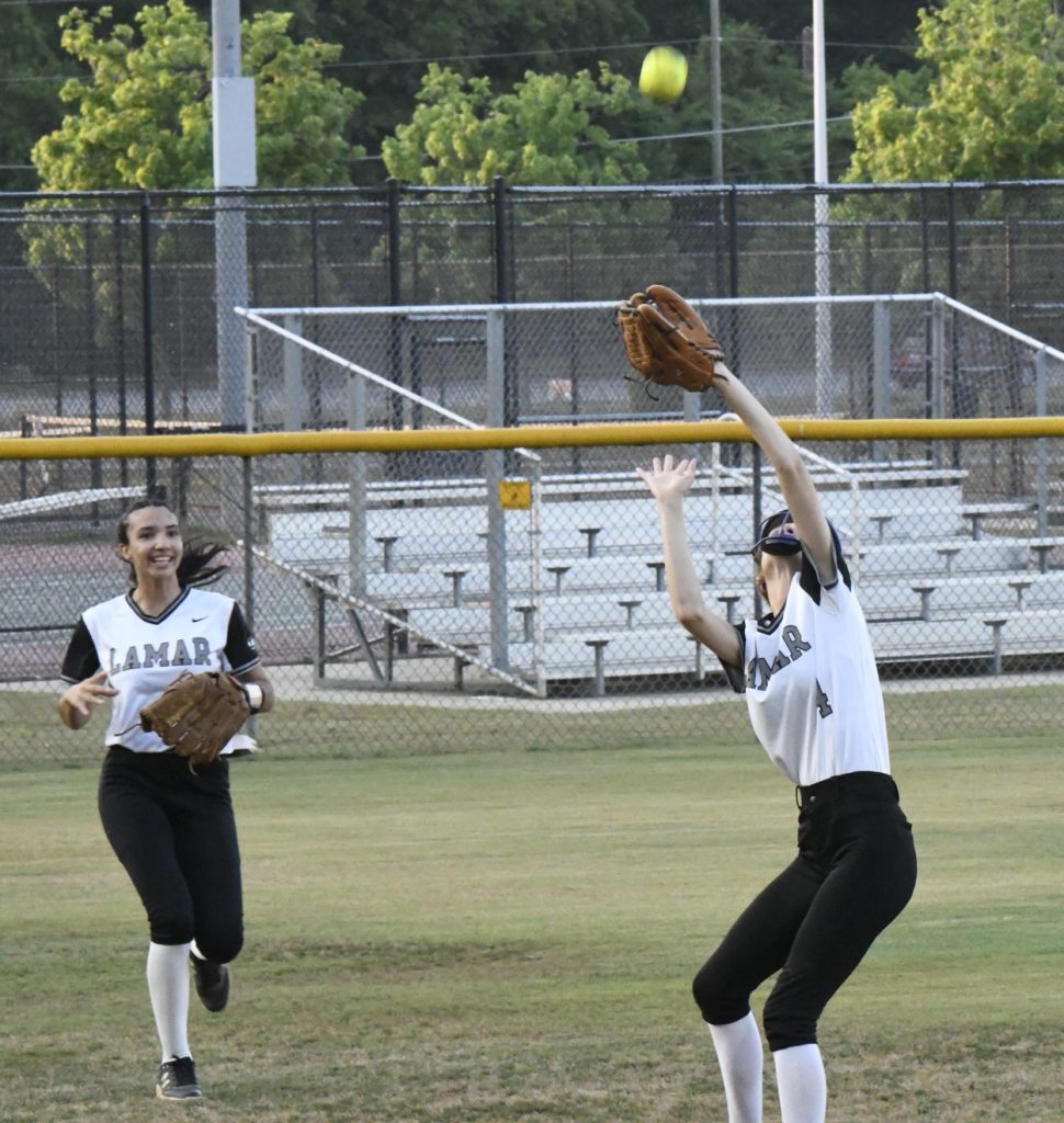Two softball players in Lamar uniforms attempt to catch a fly ball in the outfield near a yellow fence, with empty bleachers in the background.