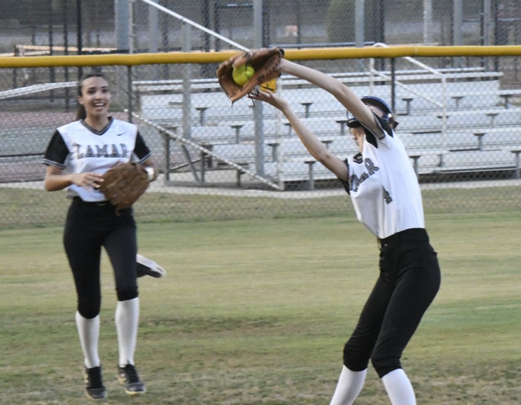 Two softball players in Lamar uniforms, one catching a ball with a glove while the other smiles nearby on a grassy field with bleachers in the background.