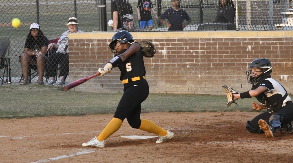 A softball player in a black uniform swings at a pitch while a catcher crouches behind home plate; spectators watch from the stands.