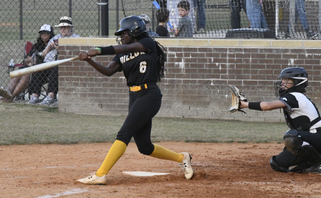 A softball player in black and yellow uniform swings at a pitch while the catcher in black and white gear prepares to catch the ball. Spectators watch behind a fence.
