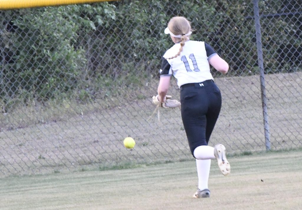 Softball player wearing number 11 in a white and black uniform runs toward a yellow softball near a chain-link fence in the outfield.